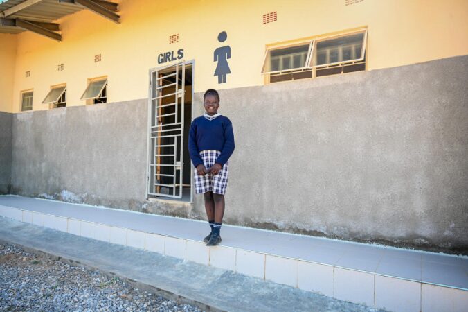 A student wearing a blue uniform standing in front of a school washroom labeled 'GIRLS'.