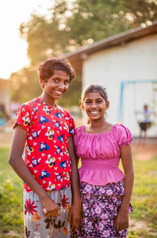 Two girls wearing colourful outfits are standing outside a school yard.