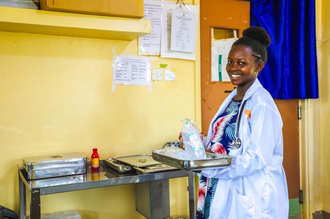 A woman in a white lab coat holding a tray in a medical room with supplies and yellow walls.
