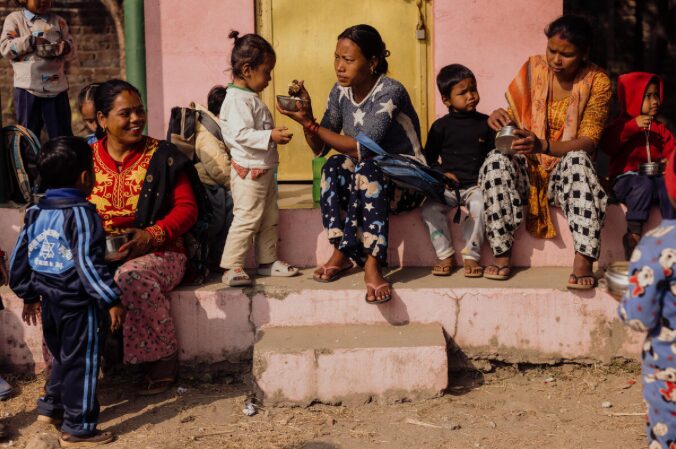 Mothers feed children sitting on a step outdoors, against a soft pink wall with a yellow door.