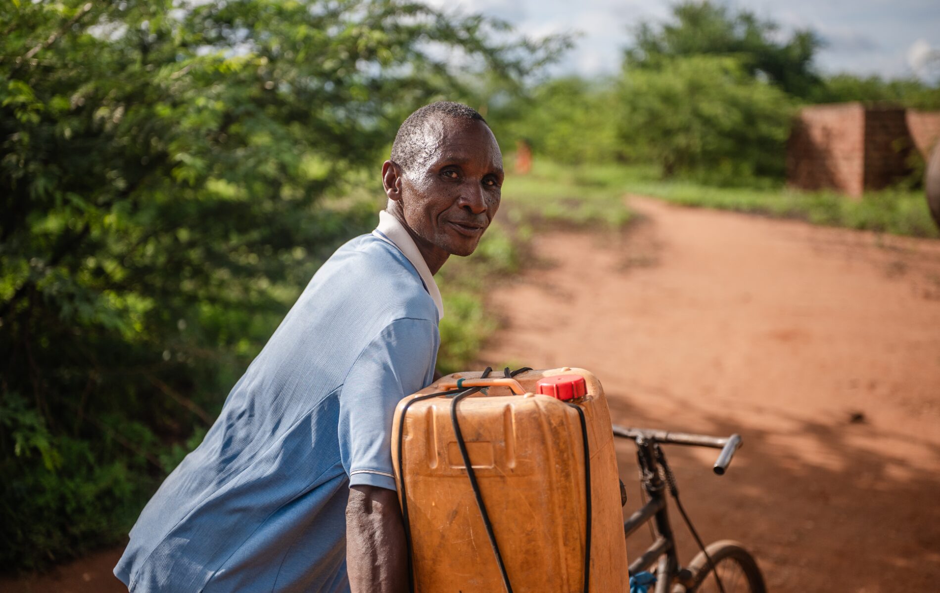 A man on a dirt road looks over his shoulder, as he balances a jerry can strapped to a bike.