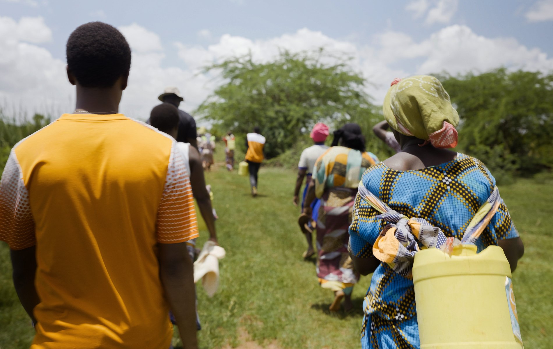 A group of men and women walk across an open field carrying buckets and jerry cans.
