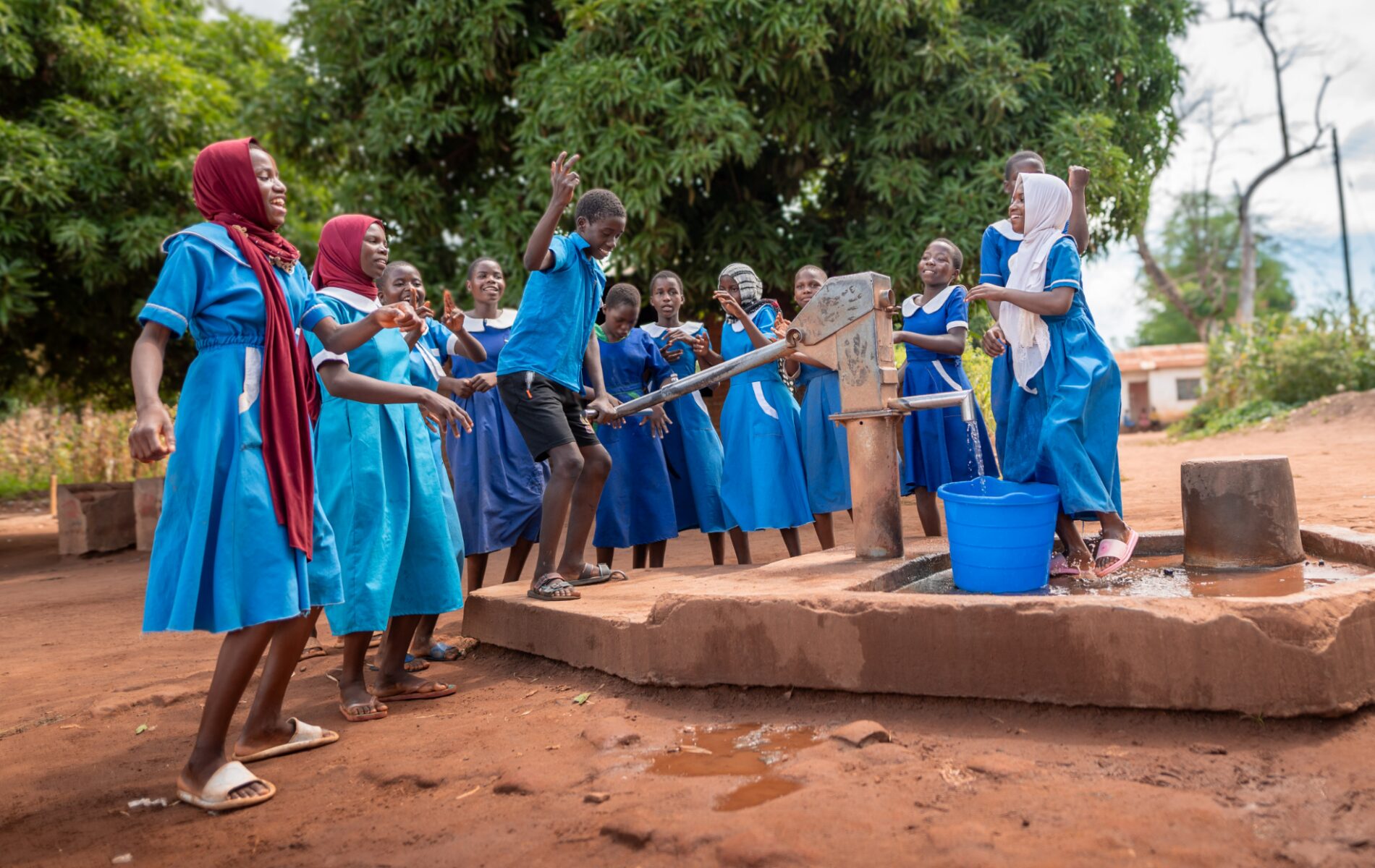 At a new community water station, a schoolboy playfully pumps water into a blue bucket as schoolgirls in blue uniforms cheer him on.