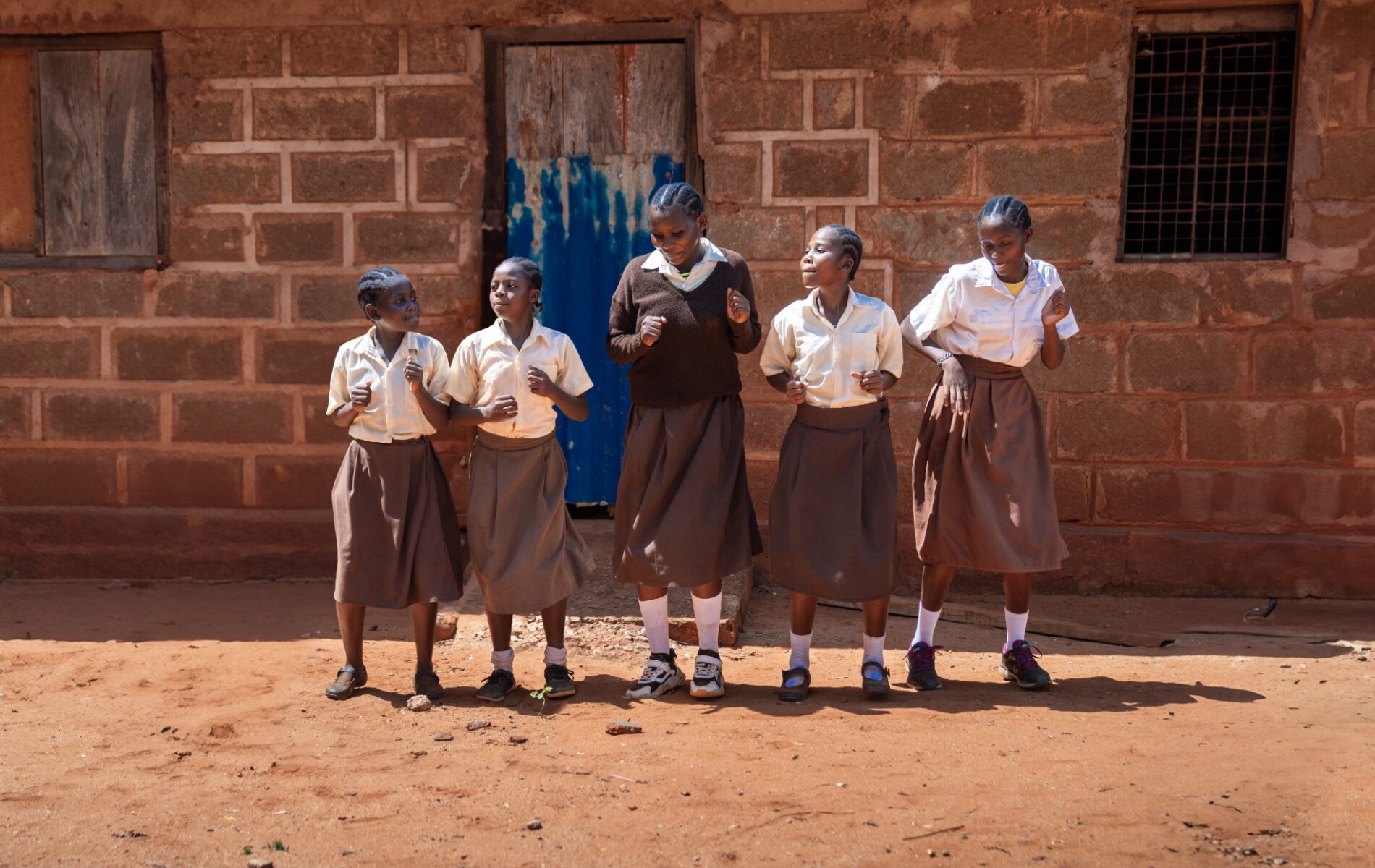 Five girls wearing brown and white school uniforms dance in front of a wall.
