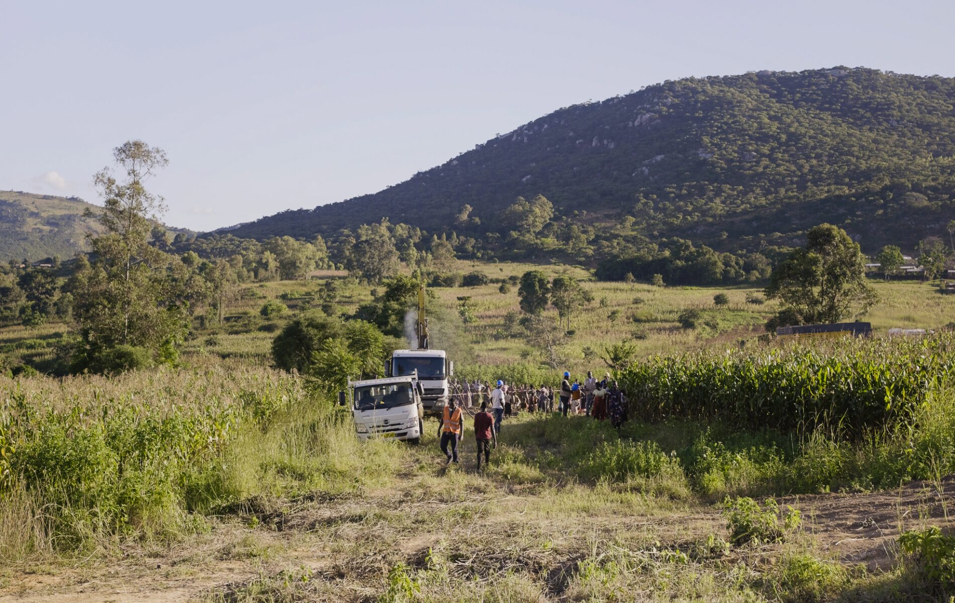 Two trucks and a group of people travel through a grassy terrain, with a mountain in the distance.