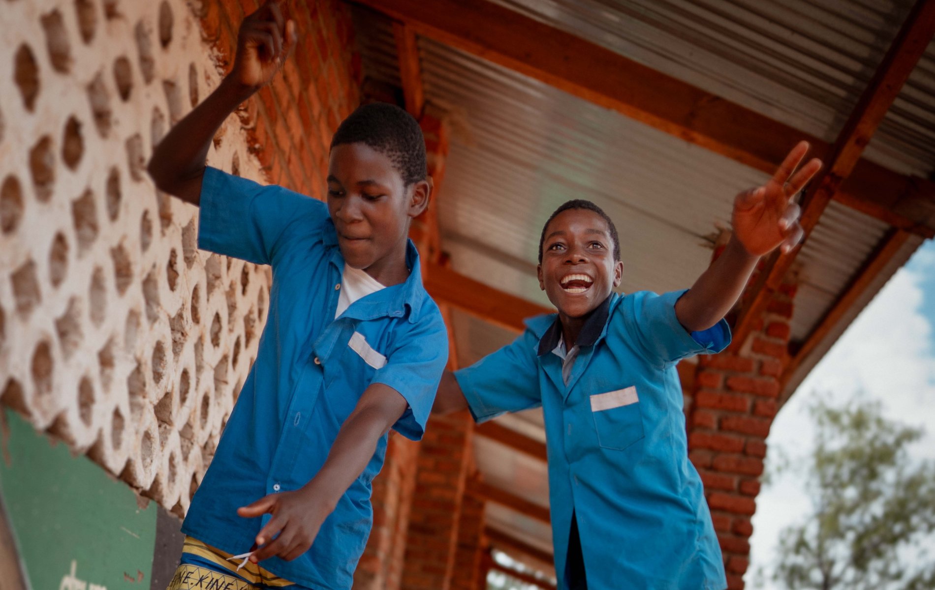Two school boys show off their dance moves outside a building with a metal roof.
