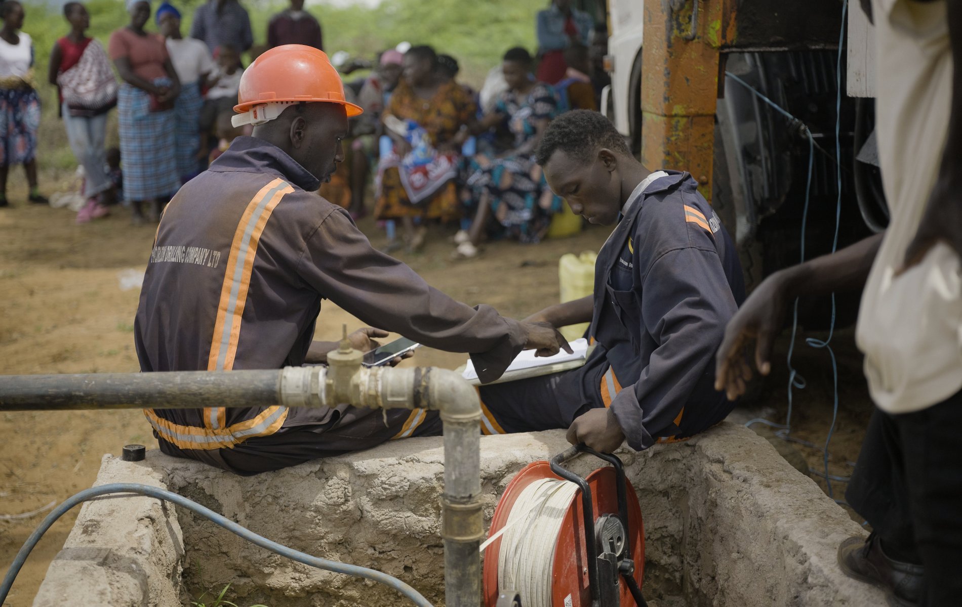 Two workers survey a borehole dig site while community members watch in the background.