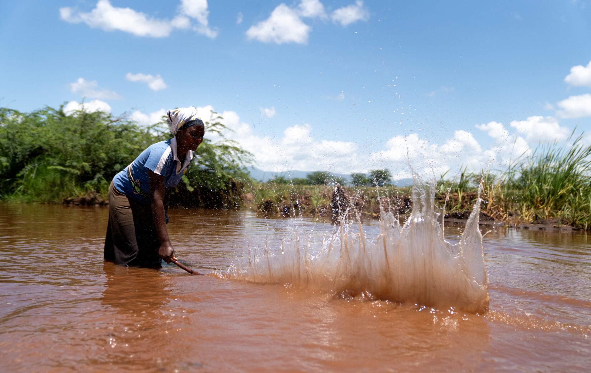 A woman uses a long stick to strike the surface of a pool of brown water, causing a big splash.