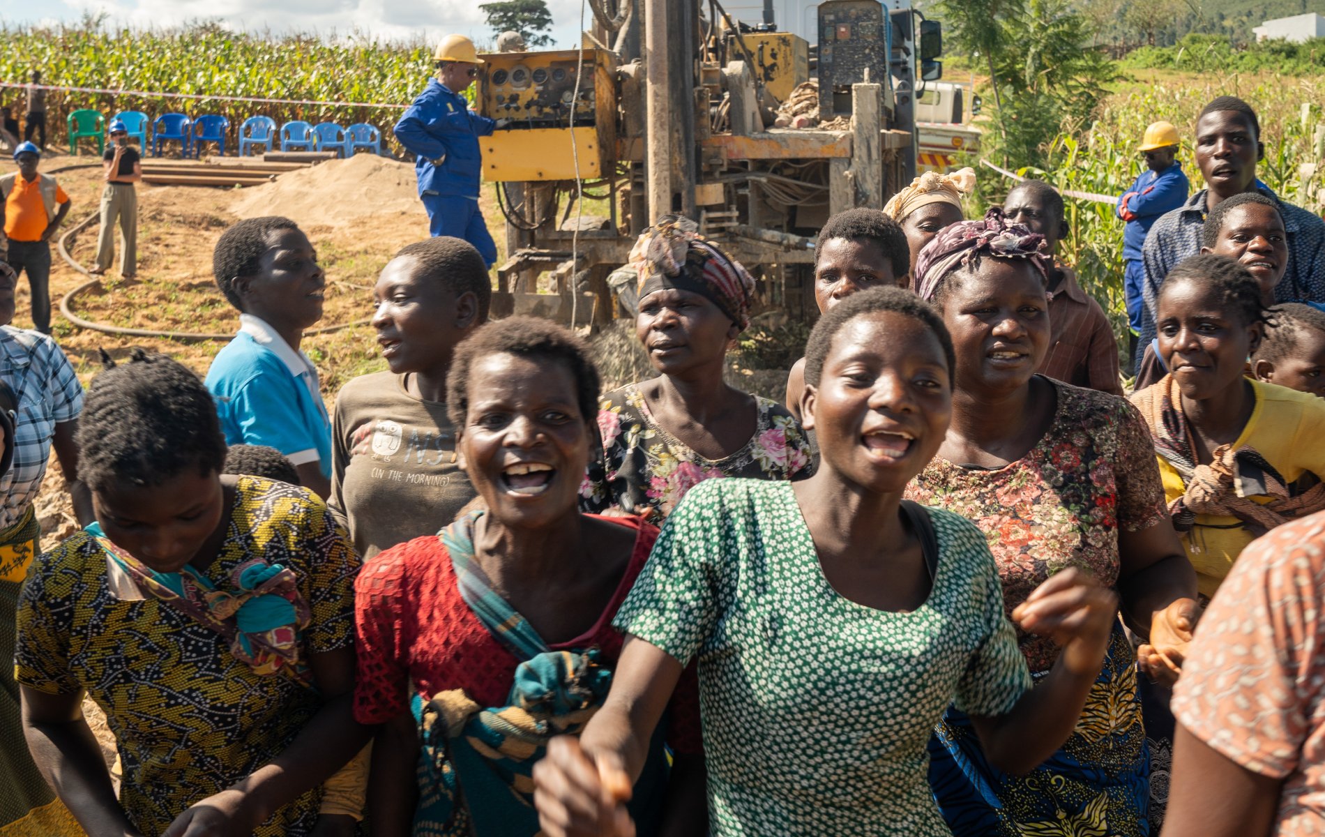 A group of women wearing colourful dresses sing and dance at the borehole dig site as workers watch.