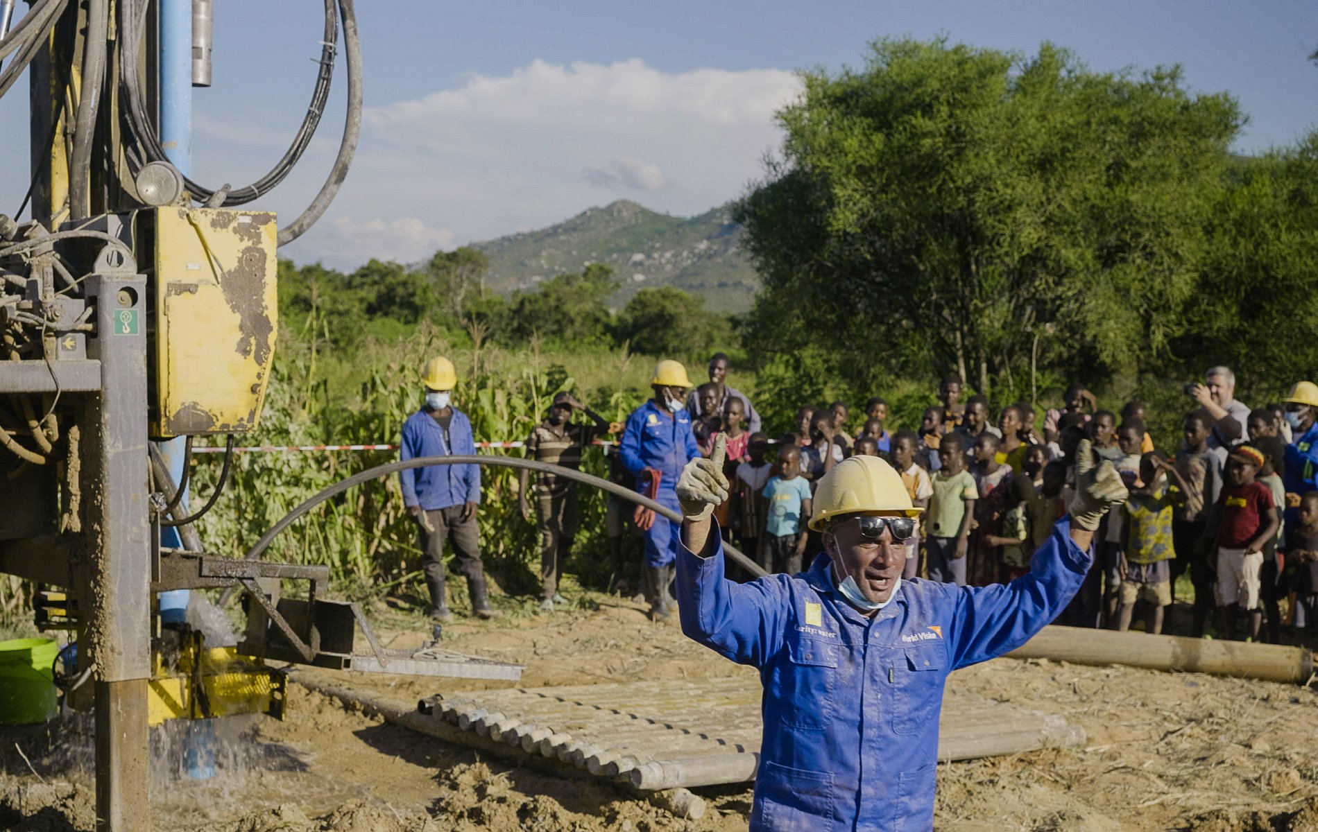 A worker wearing blue coveralls and a yellow hard hat gives two thumbs up, while a crowd watches a safe distance away.