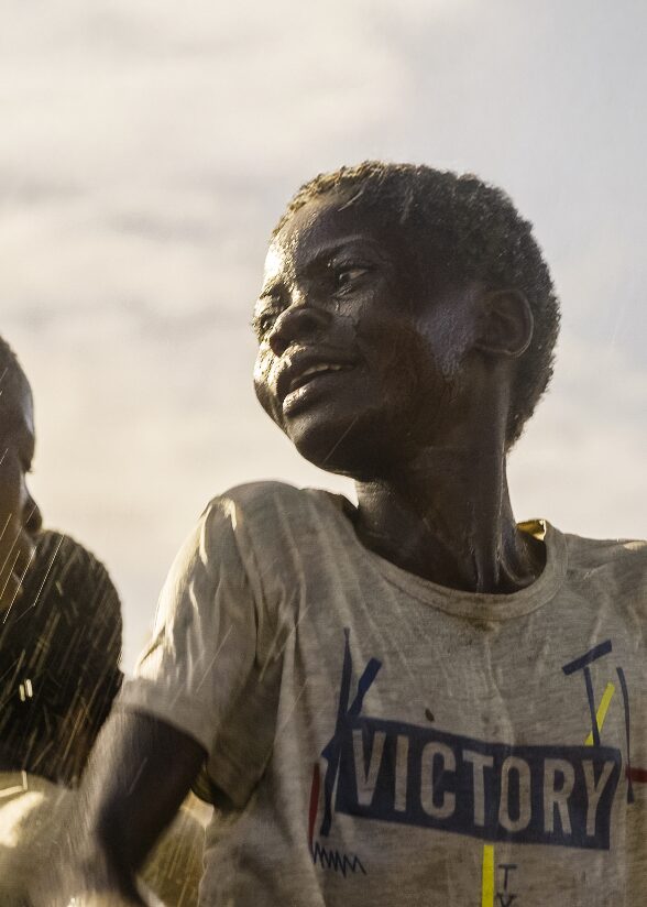 A child gazes off to the side as drops of water soak his white “Victory” t-shirt.